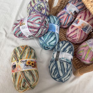 May include: A collection of Opal Aquarell yarn skeins in various colors, including blue, green, pink, and yellow. The yarn is arranged in a wicker basket and on a white surface. Each skein is labeled with the Opal Aquarell brand and product information.