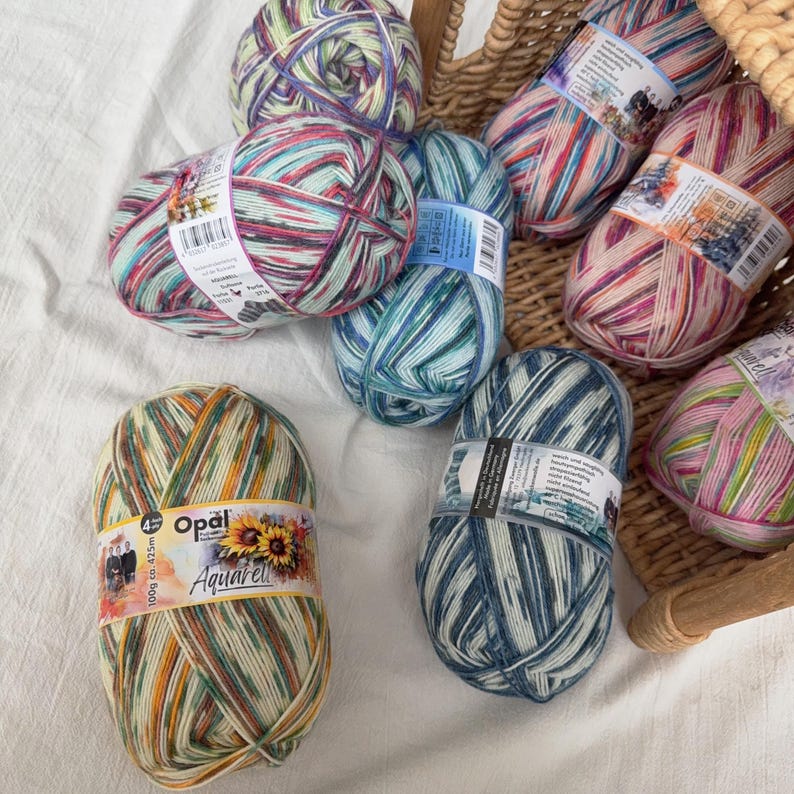 May include: A collection of Opal Aquarell yarn skeins in various colors, including blue, green, pink, and yellow. The yarn is arranged in a wicker basket and on a white surface. Each skein is labeled with the Opal Aquarell brand and product information.