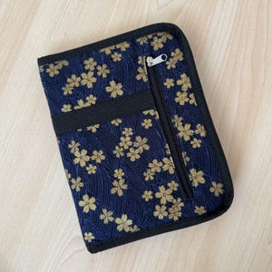 Pu&ograve; includere: Un astuccio rettangolare in tessuto con sfondo blu scuro, decorato da un motivo ripetuto di fiori di ciliegio dorati e una trama a linee ondulate. L'astuccio presenta un bordo nero, una tasca verticale con cerniera e un cursore argentato.