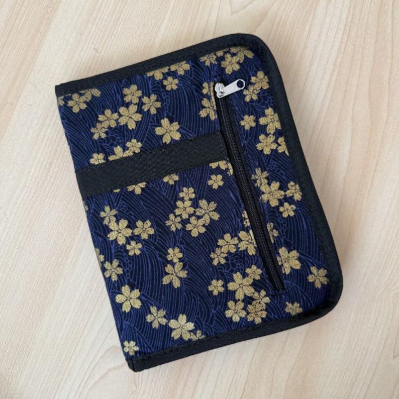 Pu&ograve; includere: Un astuccio rettangolare in tessuto con sfondo blu scuro, decorato da un motivo ripetuto di fiori di ciliegio dorati e una trama a linee ondulate. L'astuccio presenta un bordo nero, una tasca verticale con cerniera e un cursore argentato.