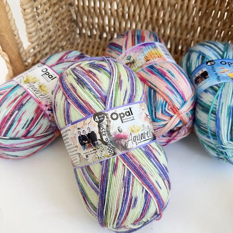 May include: Several skeins of Opal Aquarelle yarn in various colors, including green, purple, pink, and blue. The yarn is wrapped with labels that include the brand name and product details. The skeins are arranged near a woven basket.