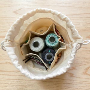 May include: A cream-colored fabric drawstring bag with an open top, featuring multiple interior pockets. Inside, the bag contains three spools of thread in light blue, dark blue, and silver, along with several metal sewing needles and small tools.