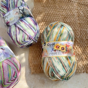 May include: Three skeins of Opal Aquarell yarn in various color combinations. The yarn is striped with shades of green, purple, yellow, and pink. Each skein has a label with the Opal logo and the word "Aquarell". The yarn is 100g.