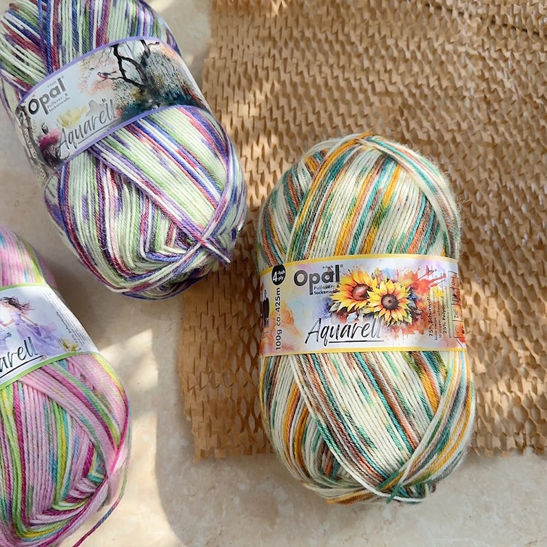 May include: Three skeins of Opal Aquarell yarn in various color combinations. The yarn is striped with shades of green, purple, yellow, and pink. Each skein has a label with the Opal logo and the word "Aquarell". The yarn is 100g.