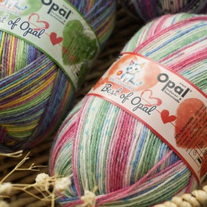 May include: Close-up of two colourful yarn skeins. The yarn is labelled "Opal Best of Opal" and features a mix of pink, green, blue, and yellow hues. The yarn is in a woven basket, with a few white flowers in the foreground.