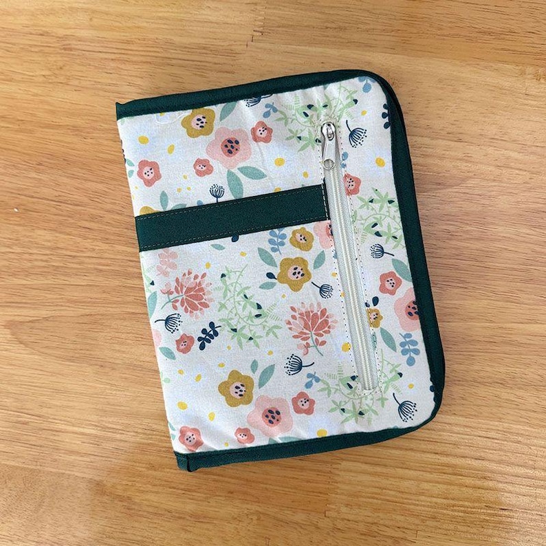Pu&ograve; includere: Una custodia organizer rettangolare in tessuto con sfondo bianco e un motivo ripetuto di fiori e foglie rosa, gialli e verdi. La custodia presenta un bordo in tessuto verde scuro e una tasca verticale con cerniera sul pannello frontale.