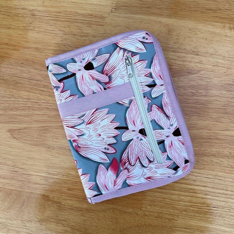 Pu&ograve; includere: Una custodia organizer rettangolare in tessuto con sfondo grigio e un motivo ripetuto di fiori di loto rosa e bianchi. La custodia presenta una fascia orizzontale rosa tinta unita sul davanti e una chiusura a zip verticale argentata a destra.