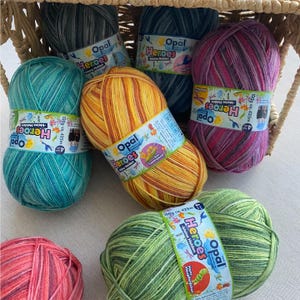 May include: A selection of colourful yarn skeins in a wicker basket. The yarn is labelled "Opal Heroes" and features shades of teal, yellow, pink, and green. Each skein weighs approximately 100g and has a length of 425m.