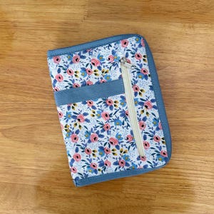 Pu&ograve; includere: Un astuccio rettangolare in tessuto con sfondo bianco e un motivo ripetuto di rose rosa e foglie blu. L'astuccio presenta un bordo in tessuto blu a tinta unita, una fascia orizzontale in tessuto blu e una cerniera color crema.