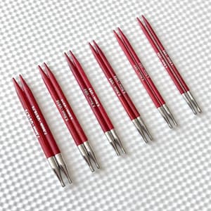 Könnte beinhalten: Ein Satz roter Stricknadeln mit silbernen Spitzen, diagonal angeordnet. Die Nadeln sind mit Größen von 4,0 mm bis 7,0 mm, US-Größen 6 bis 10,75, beschriftet. Die Nadeln bestehen aus Holz und Metall.