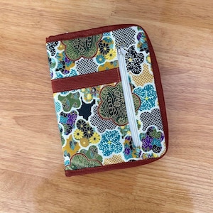 Pu&ograve; includere: Un astuccio rettangolare in tessuto con un motivo floreale e geometrico multicolore nei toni del verde, giallo, blu e nero. L'astuccio presenta un bordo in tessuto rosso scuro, una fascia orizzontale e una tasca verticale con zip bianca.
