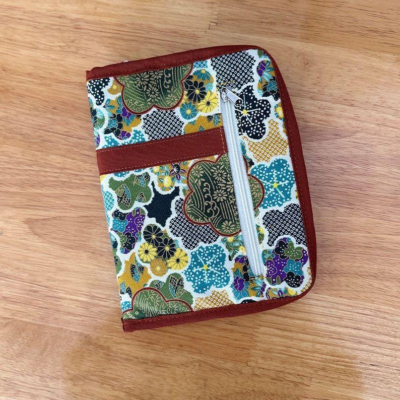 Pu&ograve; includere: Un astuccio rettangolare in tessuto con un motivo floreale e geometrico multicolore nei toni del verde, giallo, blu e nero. L'astuccio presenta un bordo in tessuto rosso scuro, una fascia orizzontale e una tasca verticale con zip bianca.