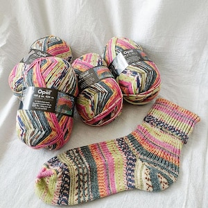 May include: A collection of Opal yarn balls and a knitted sock. The yarn is multi-colored with pink, green, yellow, and blue hues. The sock displays a similar color palette with patterned sections. The yarn balls are labeled with the brand name and product details.