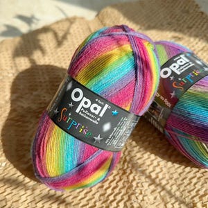 May include: Two skeins of rainbow-coloured yarn, with shades of pink, yellow, blue, and purple. The label reads "Opal Surprise" and "4-fach Pullover & Sockenwolle". Each skein weighs 100g and is approximately 425m long.