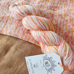 May include: A skein of hand-dyed yarn in shades of white, pink, and orange. The yarn is next to a knitted swatch with similar colors. A tag with the brand name "INTI" is attached to the yarn.