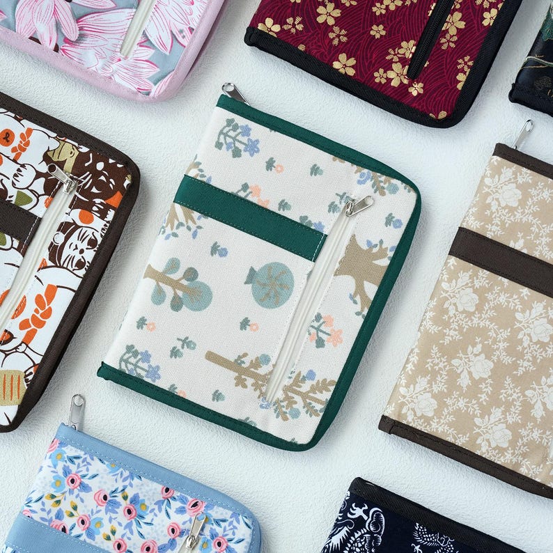 Pu&ograve; includere: Una custodia rettangolare in tessuto con base bianca caratterizzata da un motivo di piccoli alberi e fiori in tonalit&agrave; tenui di blu, verde e marrone. La custodia presenta un bordo in tessuto verde scuro, una cerniera in metallo e una tasca verticale.