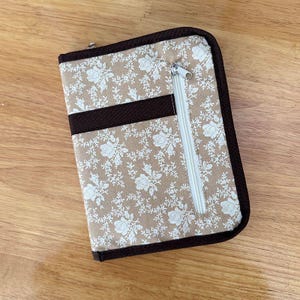 Pu&ograve; includere: Un astuccio organizer rettangolare in tessuto con sfondo beige e motivo floreale bianco. L'astuccio presenta un bordo in tessuto marrone scuro, una striscia orizzontale in tessuto marrone scuro e una tasca verticale con zip bianca sul davanti.