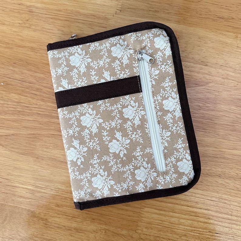 Pu&ograve; includere: Un astuccio organizer rettangolare in tessuto con sfondo beige e motivo floreale bianco. L'astuccio presenta un bordo in tessuto marrone scuro, una striscia orizzontale in tessuto marrone scuro e una tasca verticale con zip bianca sul davanti.