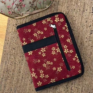 Pu&ograve; includere: Un astuccio rettangolare in tessuto con sfondo rosso intenso, un motivo ripetuto di fiori di ciliegio dorati e una trama a linee ondulate. L'astuccio presenta un bordo in tessuto nero, un nastro nero orizzontale e una tasca verticale con cerniera nera.