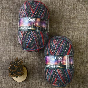 May include: Two balls of Opal Feuerwerk yarn, with a mix of red, blue, and green colours. Each ball has a label with the Opal logo and the word "FEUERWERK". A small pinecone sits on a wooden slice.