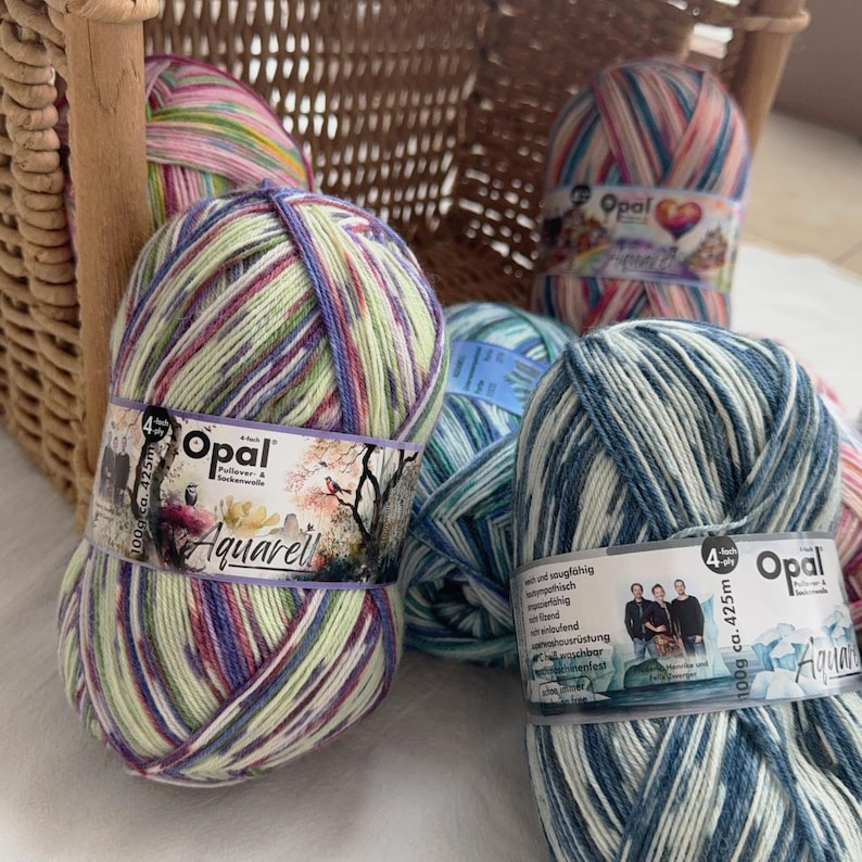 May include: Several skeins of Opal Aquarell yarn in various colors, including green, purple, and blue, are displayed. Each skein is wrapped with a label that includes the brand name and product details. The yarn is 100g and approximately 425m.