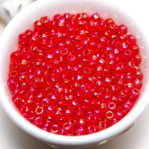 May include: A white bowl overflowing with small, translucent red beads. The beads have a glossy finish and reflect light, creating a shimmering effect. The beads are round and appear to be uniform in size.