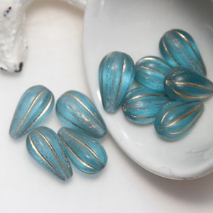 Peut inclure: Un ensemble de perles bleues claires en forme de goutte d'eau avec des rayures dorées. Les perles sont présentées dans un plat blanc en forme de cœur. Les perles ont un aspect texturé et sont probablement destinées à la création de bijoux.