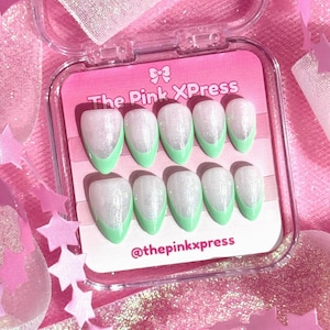 May include: A set of ten almond-shaped press-on nails in a clear plastic case. Each nail features a white glitter base with a solid light green French tip. The case insert displays the text The Pink XPress and the social media handle @thepinkxpress.