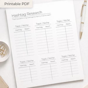 May include: A white printable PDF titled "Hashtag Research" with sections for tracking hashtags by topic. The document includes fields for "Topic/Niche," "Hashtag," and "Reach." A pen and keyboard are visible in the background.