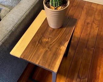 May include: A rectangular wooden side table with a two-tone design. The table features a light-colored wood section on one side and a darker, stained wood section on the other. The table is supported by black metal legs. A potted plant sits on top.