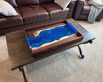 May include: A rectangular wooden serving tray with a blue resin river design. The tray is made of dark wood with a contrasting blue resin center, resting on a dark wooden coffee table.