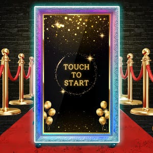 Könnte beinhalten: Ein großer, rechteckiger Fotoautomaten-Spiegel mit dekorativem Rahmen und LED-Beleuchtung. Der Spiegel zeigt den Text "TOUCH TO START" und ist von goldenen Luftballons umgeben. Die Szene umfasst einen roten Teppich und Samtseile.