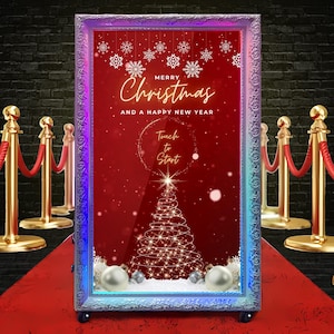 Può includere: Un grande fotomaton rettangolare a tema natalizio con sfondo rosso e cornice decorativa. Il testo "Merry Christmas and a Happy New Year" è visualizzato, insieme a un invito touch-to-start. La cabina è decorata con fiocchi di neve, un albero di Natale e ornamenti.