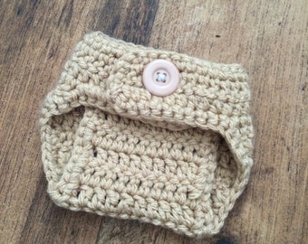 May include: A beige crochet diaper cover featuring a textured yarn construction and a single light pink button centered on the front waistband. The garment is crafted with a consistent stitch pattern and has leg openings on both sides for a secure fit.