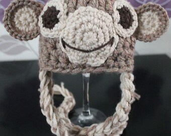 May include: A crocheted hat designed to resemble a monkey, featuring a light brown base, a cream-colored muzzle, dark brown eyes, and two large circular ears on the sides. The hat includes two long, thick braided ties hanging down from the bottom.