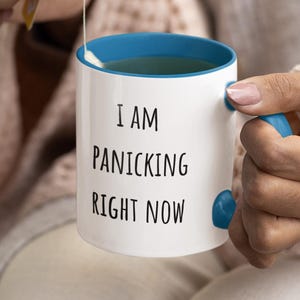 Puede incluir: Taza de cerámica blanca con interior y asa azules. La taza tiene el texto "I AM PANICKING RIGHT NOW" en negro. Una persona sostiene la taza con una bolsita de té.