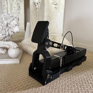 May include: A black, mechanical pedal assembly with a textured footpad and adjustable arm. The device has a rectangular base and is connected by a black wire. The background includes a white lion statue and a beige carpet.