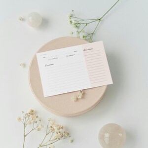 May include: A recipe card with a light pink and white design, featuring sections for title, prep time, cooking time, ingredients, and directions. The card rests on a beige circular plate, with decorative flowers and two clear spheres.
