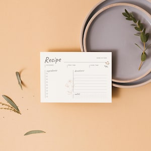 May include: A recipe card with the word "Recipe" at the top, featuring spaces for ingredients and directions. The card is cream-colored with a floral illustration. Two gray plates and sprigs of greenery are also in the image.