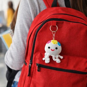Puede incluir: Un llavero de pulpo de ganchillo blanco hecho a mano con una cara sonriente y detalles de flores de colores. El llavero est&aacute; sujeto a una mochila roja, un accesorio alegre y caprichoso.