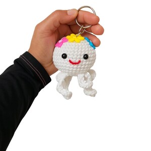 Puede incluir: Un llavero de medusa blanca de ganchillo con una cara sonriente y detalles de flores de colores. El llavero tiene un anillo de metal plateado para sujetarlo a llaves o bolsos. La medusa tiene tent&aacute;culos colgantes.