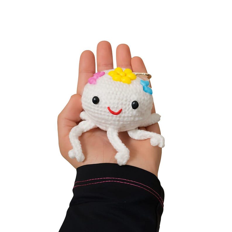 Puede incluir: Un llavero blanco con forma de medusa de ganchillo con una cara sonriente y detalles de flores de colores. El llavero tiene tent&aacute;culos colgantes y un anillo dorado. La medusa mide aproximadamente 7.6 cm de di&aacute;metro.