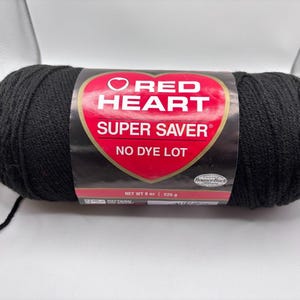 May include: A skein of black Red Heart Super Saver yarn with a red heart-shaped label. The label features the text Red Heart, Super Saver, No Dye Lot, and Net Wt 5 oz | 225 g. The yarn is wound into a cylindrical shape with a loose end visible.