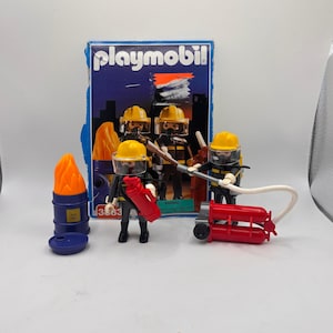 May include: A Playmobil firefighter toy set featuring two plastic figures in yellow helmets and black uniforms, a blue barrel with orange flames, a red fire extinguisher, and a red portable pump with a white hose, all in front of the original box.