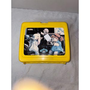 Könnte beinhalten: Eine gelbe Thermos-Lunchbox im Vintage-Stil mit einer bunten Grafik von zwei Cabbage Patch Kids-Puppen. Die Lunchbox hat eine rechteckige Form mit einem Klappdeckel und einem Griff. Das Wort "THERMOS" ist auf der Oberseite aufgedruckt.