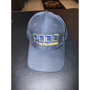 Könnte beinhalten: Marineblaue Baseballkappe mit dem Jahr "2000" in Gelb und Blau gestickt, zusammen mit dem Text "The New Millennium". Die Kappe hat einen gebogenen Schirm und zwei kleine Belüftungslöcher.