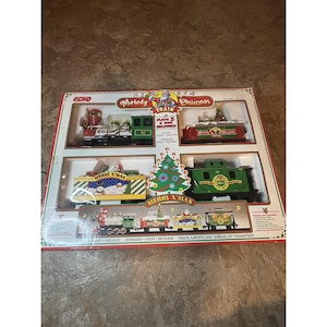 May include: A boxed Christmas train set featuring a red and green locomotive, train carriages, and a Christmas tree. The box is labelled "Melody Train Christmas" and "Merry X'mas". The track layout is a 355.6 cm circle (25.4 cm diameter).