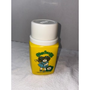 May include: A yellow Cabbage Patch Kids container with a white lid. The container features a graphic of a Cabbage Patch Kid in a blue dress. The text "Cabbage Patch Kids" is also visible.
