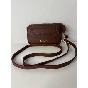 May include: A brown crossbody bag with a gold zipper and a long, adjustable strap. The bag features a decorative cut-out design on the front and the brand name "Mudd" in gold lettering.
