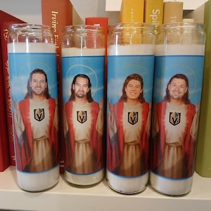 May include: Four clear glass prayer candles featuring a graphic label of a person with long hair wearing a red and white robe with a gold Vegas Golden Knights logo on the chest. Each candle has a white wax top and a light blue background with a halo.